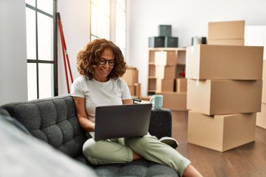 Orta yaşlı İspanyol kadın yeni evinde dizüstü bilgisayar kullanarak gülümsüyor.