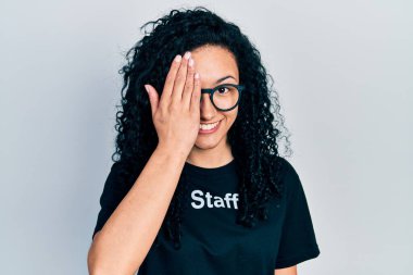Young hispanic woman with curly hair wearing staff t shirt covering one eye with hand, confident smile on face and surprise emotion. 