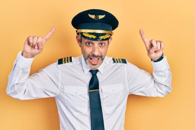 Yakışıklı, orta yaşlı, gri saçlı, uçak pilotu üniforması giyen, şaşkın, şaşkın, parmaklarıyla işaret eden ve kollarını kaldıran bir adam.. 