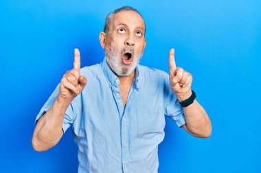 Handsome senior man with beard wearing casual blue shirt amazed and surprised looking up and pointing with fingers and raised arms. 