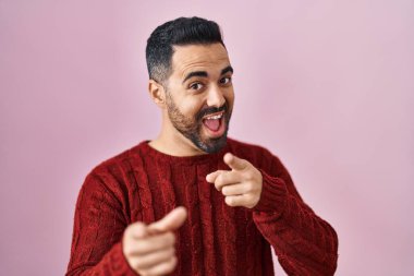 Young hispanic man with beard wearing casual sweater over pink background pointing fingers to camera with happy and funny face. good energy and vibes. 