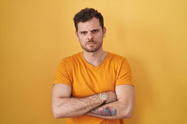 Sarı arka planda duran Latin asıllı genç adam şüpheci ve gergin, yüzünde çapraz kollu tasvip etmeyen ifade. negatif kişi. 
