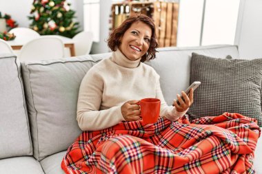 Orta yaşlı İspanyol kadın akıllı telefon kullanıyor ve evdeki Noel ağacının yanında kanepede oturup kahve içiyor..