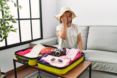 Beautiful middle age hispanic woman packing summer clothes in suitcase covering one eye with hand, confident smile on face and surprise emotion. 
