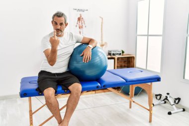 Middle age hispanic man at pain recovery clinic holding pilates ball showing middle finger, impolite and rude fuck off expression 