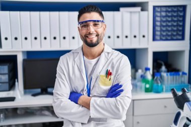 Genç Arap bilim adamı gülümsüyor. Kollarını kavuşturup laboratuvarda duruyor.