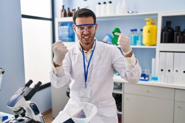Young hispanic man with beard working at scientist laboratory holding green ribbon annoyed and frustrated shouting with anger, yelling crazy with anger and hand raised 