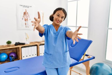 Ağrı kesici kliniğindeki güzel İspanyol fizyoterapist kadın kucaklanmak için kollarını açmış kameraya bakıyor. Mutluluğu kucaklayan neşeli bir ifade.. 