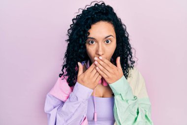 Young hispanic woman with curly hair wearing gym clothes and using headphones shocked covering mouth with hands for mistake. secret concept. 