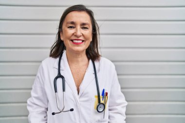 Middle age hispanic woman wearing doctor uniform and stethoscope looking positive and happy standing and smiling with a confident smile showing teeth 