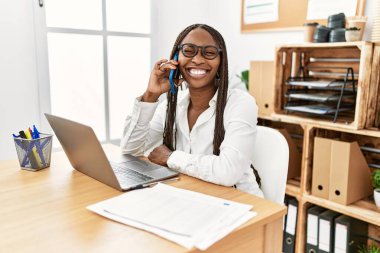 Black woman with braids working at the office speaking on the phone winking looking at the camera with sexy expression, cheerful and happy face. 