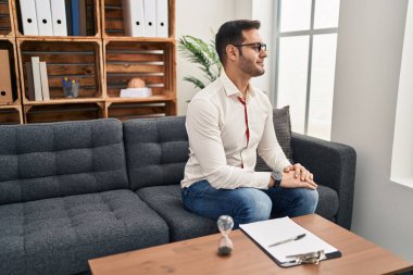 Young hispanic man with beard working at consultation office looking to side, relax profile pose with natural face and confident smile. 