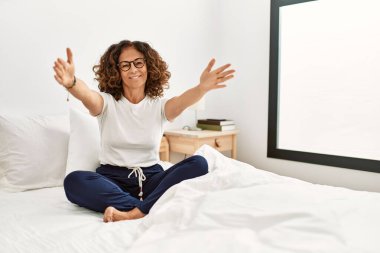 Middle age hispanic woman sitting on the bed at home looking at the camera smiling with open arms for hug. cheerful expression embracing happiness. 