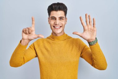 Young hispanic man standing over blue background showing and pointing up with fingers number seven while smiling confident and happy. 