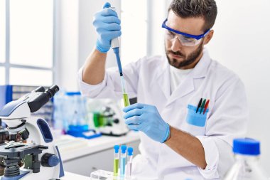 Laboratuvarda pipet ve test tüpü kullanarak bilim adamı üniforması giyen İspanyol genç bir adam.