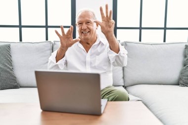 Senior man using laptop at home sitting on the sofa showing and pointing up with fingers number nine while smiling confident and happy. 
