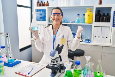 Hispanic woman working at scientist laboratory screaming proud, celebrating victory and success very excited with raised arm 