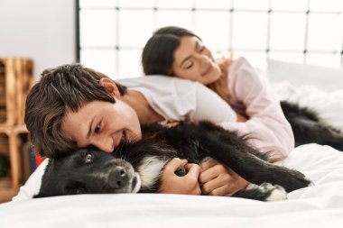 Kafkasyalı genç bir çift mutlu bir şekilde yatakta uzanıyor ve evde köpekle oynuyor..