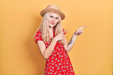 Beautiful caucasian woman with blond hair wearing summer hat smiling and looking at the camera pointing with two hands and fingers to the side. 