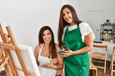 İki Latin resim öğrencisi gülümsüyor, resim yapıyor ve sanat okulunda akıllı telefon kullanıyor..