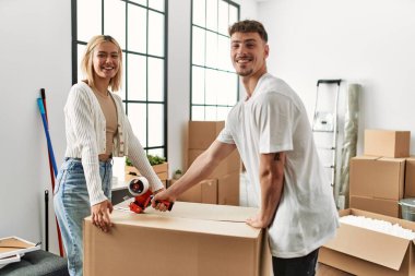 Genç beyaz çift yeni evlerinde koli bandı kullanarak mutlu bir kapanış kutusuna gülümsüyor..