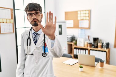 Hispanic man with beard wearing doctor uniform and stethoscope at the office doing stop sing with palm of the hand. warning expression with negative and serious gesture on the face. 