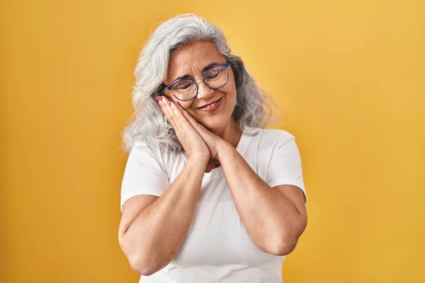 Middle age woman with grey hair standing over yellow background sleeping tired dreaming and posing with hands together while smiling with closed eyes.