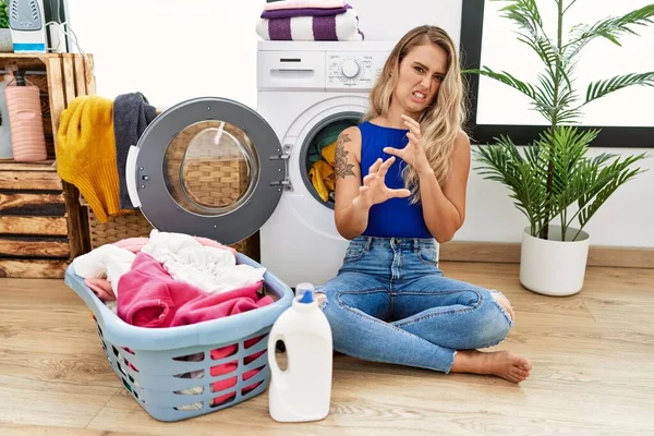 Young beautiful woman doing laundry sitting by wicker basket disgusted expression, displeased and fearful doing disgust face because aversion reaction. with hands raised