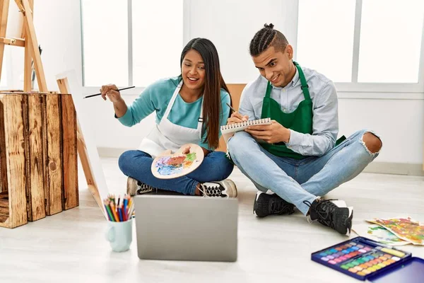 Young latin painter couple having online paint class using laptop sitting on the floor at art studio.