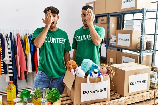 Young gay couple wearing volunteer t shirt at donations stand covering one eye with hand, confident smile on face and surprise emotion.