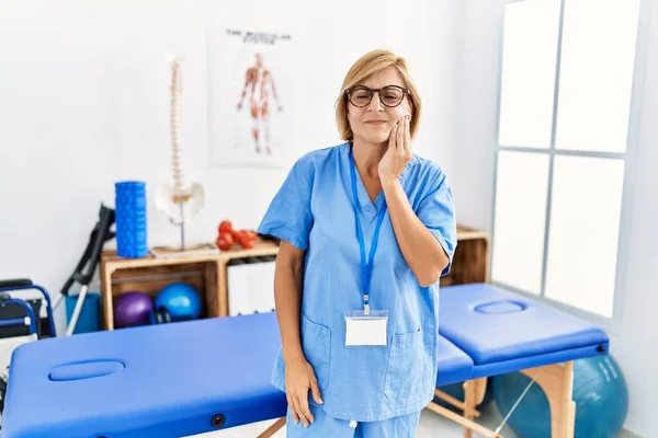 Middle age blonde woman working at pain recovery clinic touching mouth with hand with painful expression because of toothache or dental illness on teeth. dentist