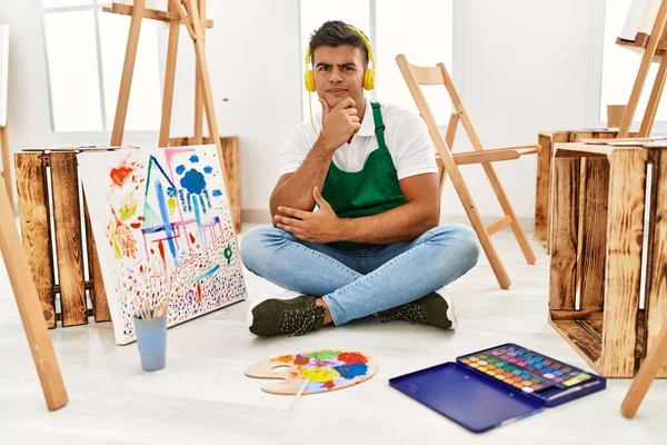 Young hispanic man at art studio looking confident at the camera with smile with crossed arms and hand raised on chin. thinking positive.