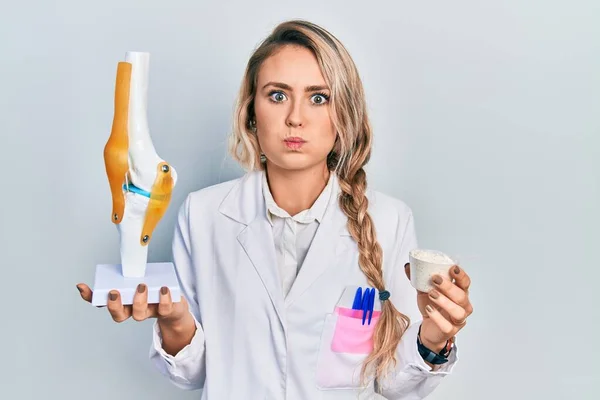 Beautiful young blonde doctor woman holding anatomical model of knee joint and protein powder puffing cheeks with funny face. mouth inflated with air, catching air.