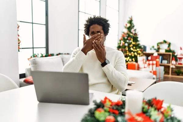 Young african american man using laptop sitting on the table by christmas tree smelling something stinky and disgusting, intolerable smell, holding breath with fingers on nose. bad smell