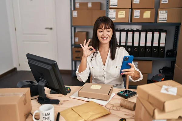 年轻的黑发女人在小企业电子商务中工作 用智能手机用手指做着确定的手势 微笑着友好的手势表示极好的象征 — 图库照片