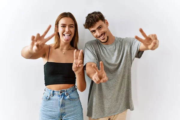 Young beautiful couple standing together over isolated background smiling with tongue out showing fingers of both hands doing victory sign. number two.
