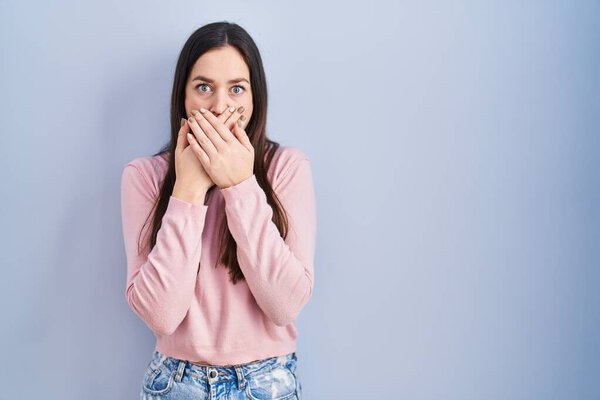 Молодая брюнетка, стоящая на синем фоне, потрясена, закрывая рот руками за ошибку. секретное понятие. 