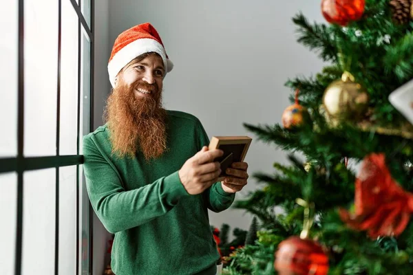Young Redhead Man Looking Photo Standing Christmas Tree Home — Φωτογραφία Αρχείου