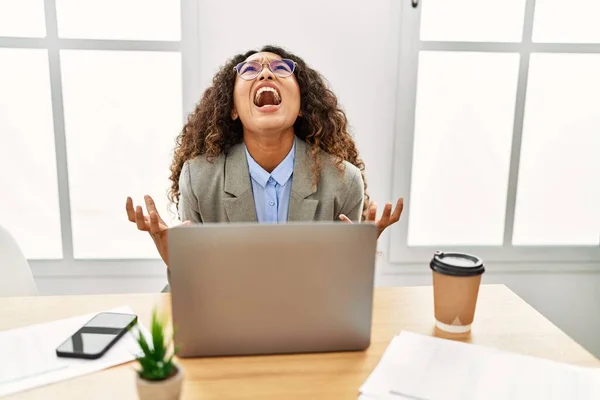 Beautiful hispanic business woman sitting on desk at office working with laptop crazy and mad shouting and yelling with aggressive expression and arms raised. frustration concept.
