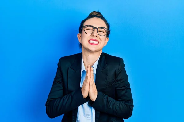 Young hispanic girl wearing business clothes and glasses begging and praying with hands together with hope expression on face very emotional and worried. begging.