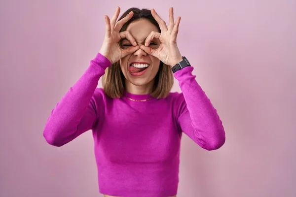 西班牙女人站在粉红的背景上做着像双筒望远镜一样的手势 伸出舌头 眼睛透过手指看着 疯狂的表达 — 图库照片