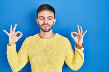 Mavi arka planda duran İspanyol adam rahatlamış ve gözleri kapalı gülümsüyor parmaklarıyla meditasyon hareketi yapıyor. yoga konsepti. 
