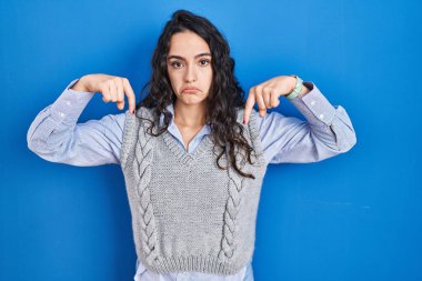 Mavi arka planda duran esmer kadın üzgün ve üzgün görünüyor, parmaklarıyla yönü gösteriyor, mutsuz ve depresif.. 