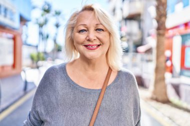 Orta yaşlı sarışın kadın sokakta kendinden emin bir şekilde gülümsüyor.