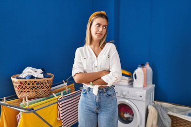 Young blonde woman at laundry room looking to the side with arms crossed convinced and confident 
