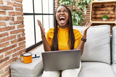 African woman sitting on the sofa using laptop at home crazy and mad shouting and yelling with aggressive expression and arms raised. frustration concept. 