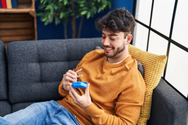 Young hispanic man holding invisible aligner sitting on sofa at home