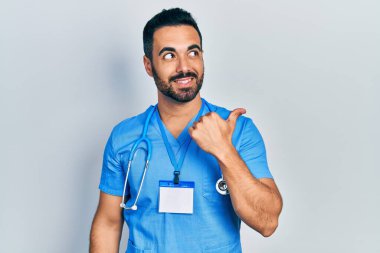 Yakışıklı, sakallı, doktor üniforması giyen mutlu yüzüyle gülümseyen ve baş parmağıyla yüzünü gösteren bir İspanyol.. 