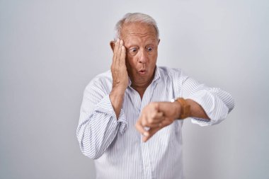 Senior man with grey hair standing over isolated background looking at the watch time worried, afraid of getting late 
