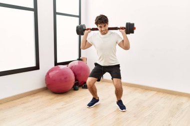 Genç İspanyol adam gülümsüyor. Spor merkezinde halterle antrenman yapıyor.